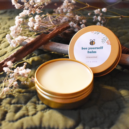 An open, gold tin of unscented balm labeled 'Bee Yourself Balm' for lip and skin care, placed on a green quilted background. Dried flowers and wooden sticks surround the balm, creating a natural and rustic aesthetic.