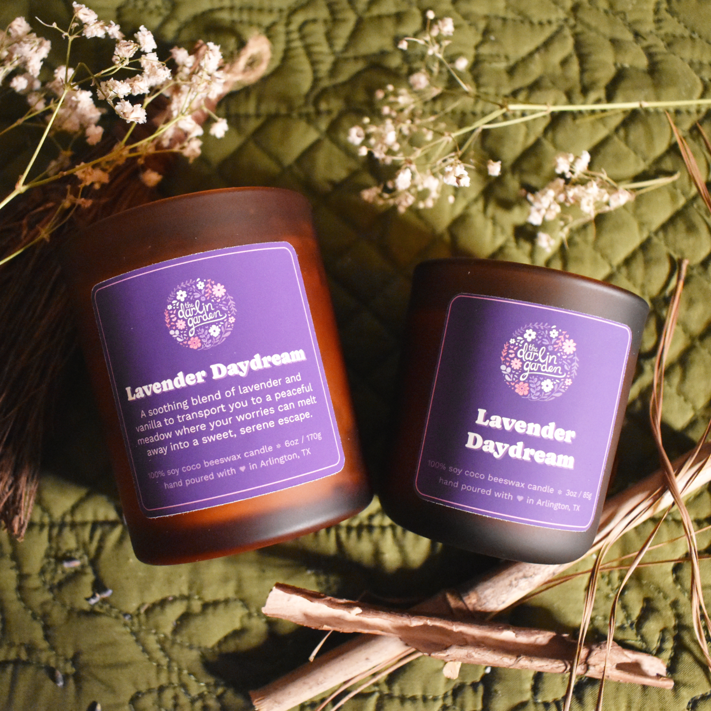 Two amber glass candles labeled 'Lavender Daydream' from The Darlin' Garden, displayed on a quilted green background with dried flowers and wooden sticks. The candles features a soothing blend of lavender and vanilla, emphasizing its natural and serene qualities.